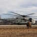 A U.S. Marines heavy-lift helicopter, identified as a Sikorsky CH-53E Super Stallion marked 'MARINES' with tail number 23, is on or near a tarmac with rotors spinning. Two Marines in combat gear are crouching nearby, appearing to wait for or prepare to board the aircraft. The setting appears to be a military training area with arid hills in the background.