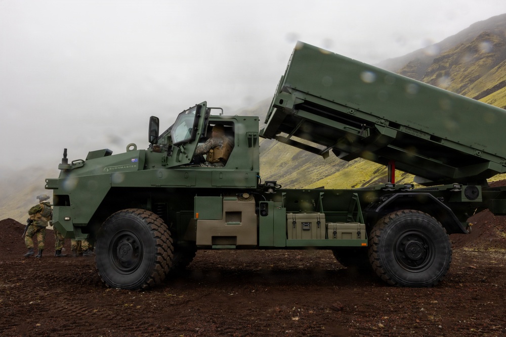 A military vehicle identified as a Bushmaster is positioned on dark volcanic terrain with a launcher system elevated at an angle. At least two soldiers in camouflage uniforms are visible near the vehicle. The setting appears to be a mountainous, overcast environment with green moss-covered hillsides in the background.