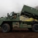 A military vehicle identified as a Bushmaster is positioned on dark volcanic terrain with a launcher system elevated at an angle. At least two soldiers in camouflage uniforms are visible near the vehicle. The setting appears to be a mountainous, overcast environment with green moss-covered hillsides in the background.