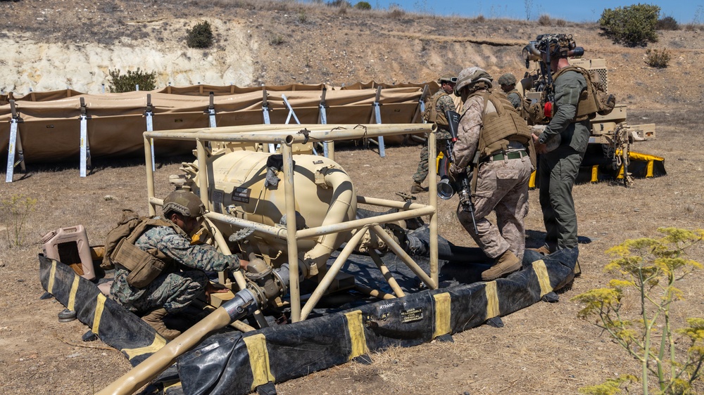 Military personnel in camouflage uniforms are working around a tan-colored fuel or water bladder/tank system enclosed in a metal frame, surrounded by a black and yellow spill containment berm. The scene appears to be a field training exercise at an arid outdoor location, with additional tan shelter structures visible in the background. A civilian or contractor with a camera system is also present observing or documenting the activity.