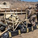 Military personnel in camouflage uniforms are working around a tan-colored fuel or water bladder/tank system enclosed in a metal frame, surrounded by a black and yellow spill containment berm. The scene appears to be a field training exercise at an arid outdoor location, with additional tan shelter structures visible in the background. A civilian or contractor with a camera system is also present observing or documenting the activity.