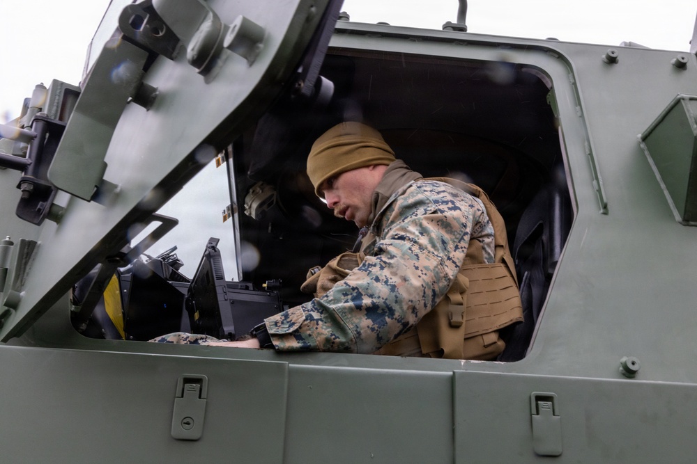 A military service member wearing MARPAT (Marine pattern) camouflage uniform and a tan beanie is leaning into the open hatch of a military armored vehicle. The individual appears to be operating or inspecting equipment inside the vehicle's cockpit/crew compartment. The vehicle is painted in olive drab green, consistent with U.S. military ground vehicles.
