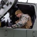 A military service member wearing MARPAT (Marine pattern) camouflage uniform and a tan beanie is leaning into the open hatch of a military armored vehicle. The individual appears to be operating or inspecting equipment inside the vehicle's cockpit/crew compartment. The vehicle is painted in olive drab green, consistent with U.S. military ground vehicles.
