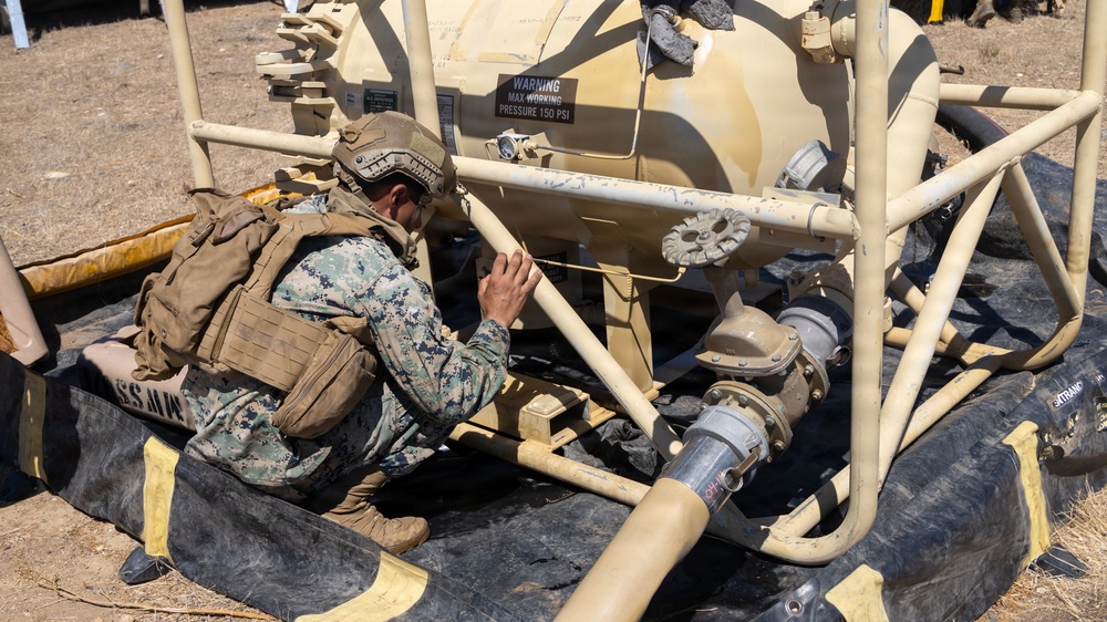A military service member in digital camouflage uniform and tactical gear is crouching down to inspect or work on a tan-colored pressurized tank system with piping and valves. The equipment appears to be a portable fluid storage and distribution unit, likely used for water or fuel operations. The setting appears to be an outdoor field environment.