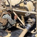 A military service member in digital camouflage uniform and tactical gear is crouching down to inspect or work on a tan-colored pressurized tank system with piping and valves. The equipment appears to be a portable fluid storage and distribution unit, likely used for water or fuel operations. The setting appears to be an outdoor field environment.