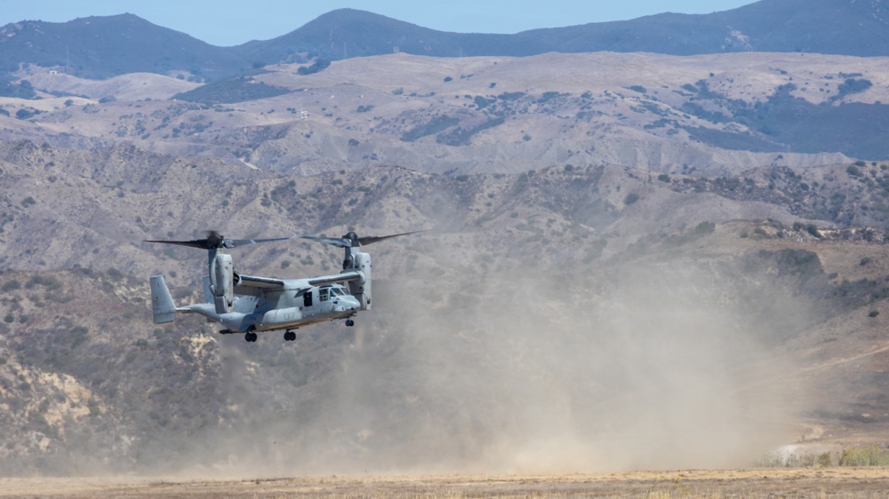 A military tiltrotor aircraft, identifiable as a Bell Boeing V-22 Osprey, is captured in a low-altitude hover or landing phase over an arid, open field. The aircraft's rotors are tilted upward in helicopter mode, kicking up a large dust cloud beneath it. The backdrop features dry, rolling hills and mountains consistent with a Southern California or similar semi-arid training environment.