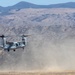A military tiltrotor aircraft, identifiable as a Bell Boeing V-22 Osprey, is captured in a low-altitude hover or landing phase over an arid, open field. The aircraft's rotors are tilted upward in helicopter mode, kicking up a large dust cloud beneath it. The backdrop features dry, rolling hills and mountains consistent with a Southern California or similar semi-arid training environment.