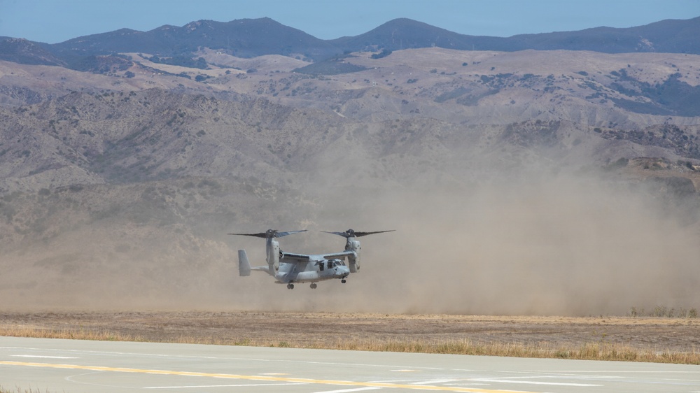 A military tiltrotor aircraft, identifiable as a V-22 Osprey, is captured hovering or landing near a runway, generating a large dust cloud behind it. The setting appears to be an arid military airfield or training area with dry, brown terrain and rugged mountains in the background. The image documents a rotorcraft operation in a desert-like environment under clear skies.