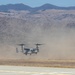 A military tiltrotor aircraft, identifiable as a V-22 Osprey, is captured hovering or landing near a runway, generating a large dust cloud behind it. The setting appears to be an arid military airfield or training area with dry, brown terrain and rugged mountains in the background. The image documents a rotorcraft operation in a desert-like environment under clear skies.