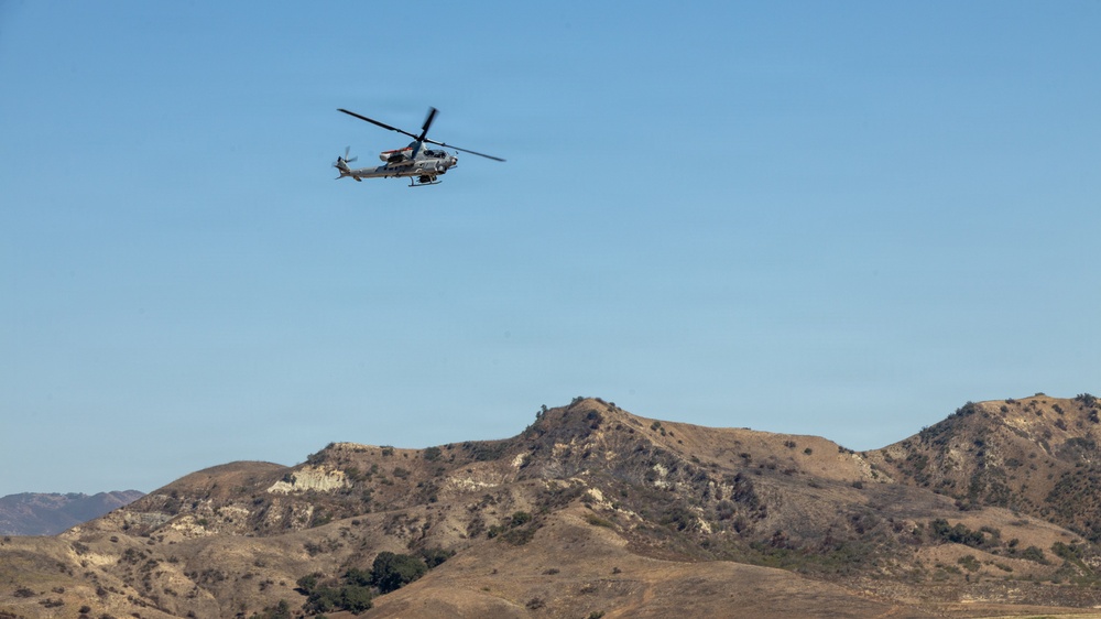 A military attack helicopter is captured in flight over a dry, arid mountainous landscape under a clear blue sky. The helicopter appears to be a tandem-seat attack variant, likely an AH-1Z Viper or similar model, flying at low-to-medium altitude. The terrain below is characterized by brown, vegetation-sparse hills typical of Southern California or similar semi-arid regions.