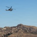 A military attack helicopter is captured in flight over a dry, arid mountainous landscape under a clear blue sky. The helicopter appears to be a tandem-seat attack variant, likely an AH-1Z Viper or similar model, flying at low-to-medium altitude. The terrain below is characterized by brown, vegetation-sparse hills typical of Southern California or similar semi-arid regions.
