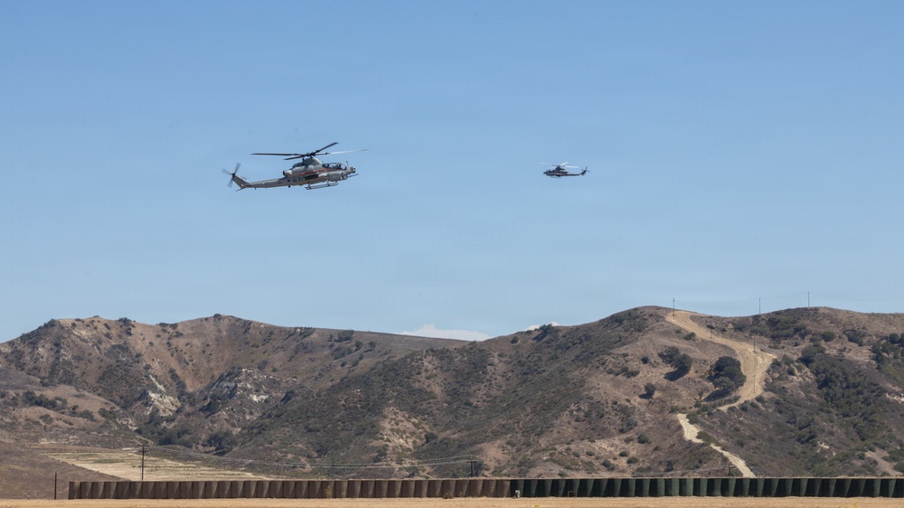 Two military helicopters, likely AH-1Z Viper attack helicopters, are flying in formation over an arid, hilly landscape. The terrain features dry, brown hills with sparse vegetation, consistent with a military training installation in Southern California. A low barrier or berm structure is visible in the foreground.