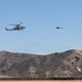 Two military helicopters, likely AH-1Z Viper attack helicopters, are flying in formation over an arid, hilly landscape. The terrain features dry, brown hills with sparse vegetation, consistent with a military training installation in Southern California. A low barrier or berm structure is visible in the foreground.