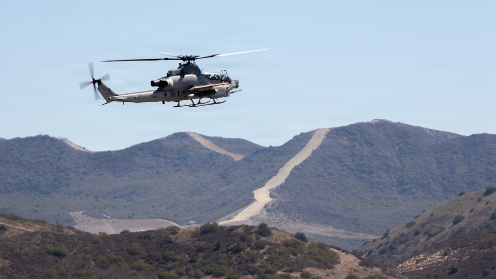 A military attack helicopter is captured in flight over a hilly, arid landscape. The aircraft appears to be a U.S. Marine Corps AH-1Z Viper, identifiable by its tandem cockpit, stub wings with weapon pylons, and skid landing gear. In the background, a dirt road or firebreak cuts across the ridgeline of scrub-covered hills under a clear sky.