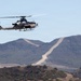 A military attack helicopter is captured in flight over a hilly, arid landscape. The aircraft appears to be a U.S. Marine Corps AH-1Z Viper, identifiable by its tandem cockpit, stub wings with weapon pylons, and skid landing gear. In the background, a dirt road or firebreak cuts across the ridgeline of scrub-covered hills under a clear sky.