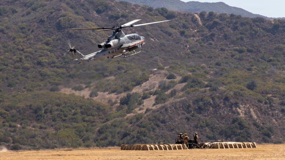 A military attack helicopter, identifiable as a Bell AH-1Z Viper, is flying low over an arid training field with hills covered in scrub vegetation in the background. On the ground below, several personnel in military uniforms are positioned near what appears to be a large inflatable or fabric ground marker/landing pad. The scene is consistent with a military training exercise at a base in a semi-arid environment.