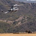 A military attack helicopter, identifiable as a Bell AH-1Z Viper, is flying low over an arid training field with hills covered in scrub vegetation in the background. On the ground below, several personnel in military uniforms are positioned near what appears to be a large inflatable or fabric ground marker/landing pad. The scene is consistent with a military training exercise at a base in a semi-arid environment.