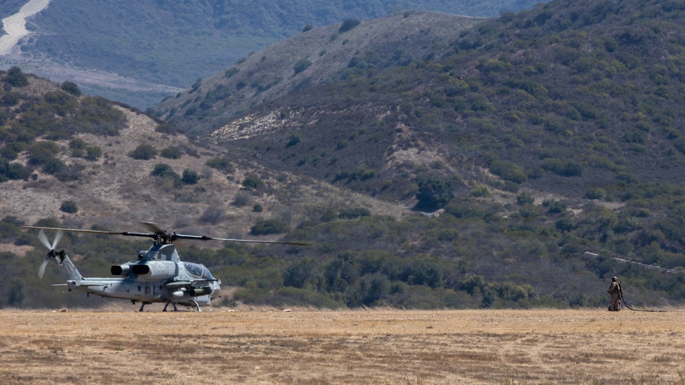 A military attack helicopter, identifiable as an AH-1Z Viper or similar Cobra variant, is on the ground in an arid, open field with rotor blades spinning. A single personnel figure stands to the right of the aircraft, appearing to hold a fuel hose or tether line. The scene is set against a backdrop of dry, scrub-covered hills consistent with a Southern California or similar semi-arid training environment.
