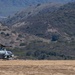 A military attack helicopter, identifiable as an AH-1Z Viper or similar Cobra variant, is on the ground in an arid, open field with rotor blades spinning. A single personnel figure stands to the right of the aircraft, appearing to hold a fuel hose or tether line. The scene is set against a backdrop of dry, scrub-covered hills consistent with a Southern California or similar semi-arid training environment.