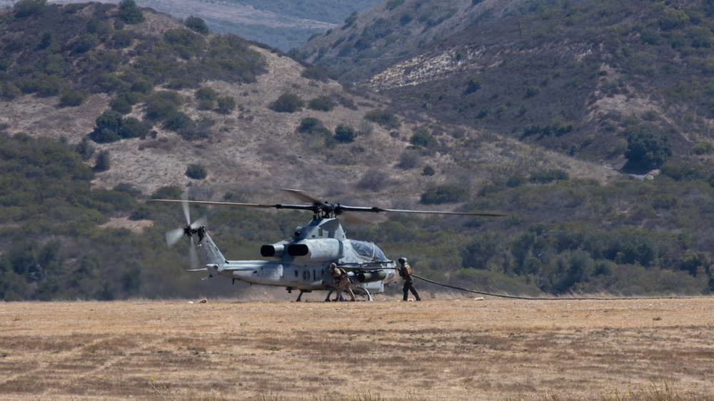A military attack helicopter, identifiable as an AH-1Z Viper based on its tandem cockpit and stub wings, is on the ground in an open dry field with personnel conducting what appears to be a forward arming and refueling point (FARP) operation. Two or three ground crew members are visible near the aircraft, one appearing to handle a fuel hose. The setting is a semi-arid valley surrounded by scrub-covered hills.