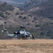 A military attack helicopter, identifiable as an AH-1Z Viper based on its tandem cockpit and stub wings, is on the ground in an open dry field with personnel conducting what appears to be a forward arming and refueling point (FARP) operation. Two or three ground crew members are visible near the aircraft, one appearing to handle a fuel hose. The setting is a semi-arid valley surrounded by scrub-covered hills.