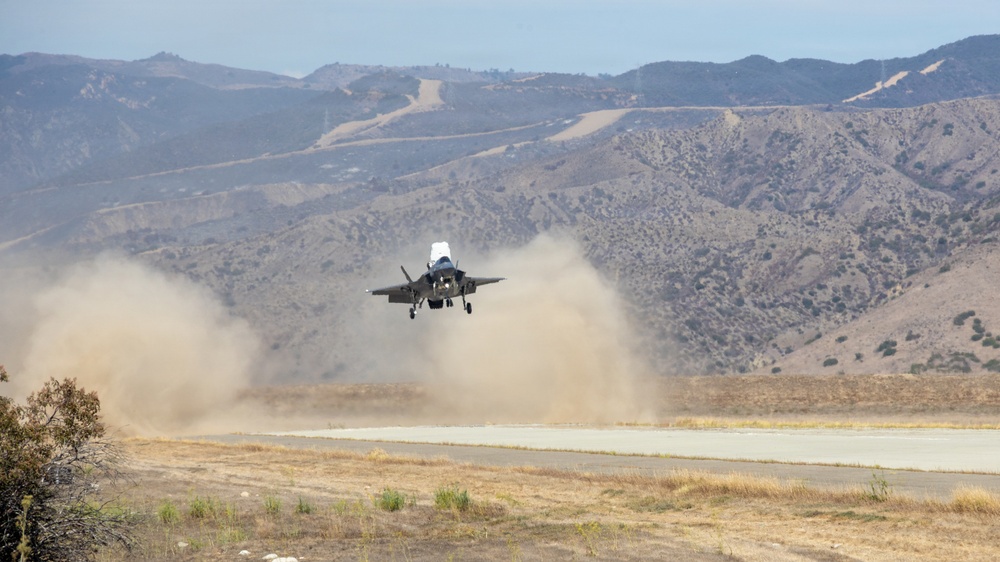 A military jet aircraft, consistent with an F-35 Lightning II, is captured during a short takeoff or vertical landing maneuver on a desert airstrip. The aircraft's powerful downwash is generating large dust clouds on either side of the runway. The setting appears to be an arid, mountainous region, likely a military training or test facility.
