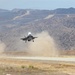 A military jet aircraft, consistent with an F-35 Lightning II, is captured during a short takeoff or vertical landing maneuver on a desert airstrip. The aircraft's powerful downwash is generating large dust clouds on either side of the runway. The setting appears to be an arid, mountainous region, likely a military training or test facility.