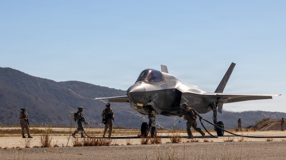 A military fifth-generation stealth fighter jet, identifiable as an F-35, is positioned on a tarmac or forward operating strip in a semi-arid, hilly environment. Ground crew personnel in combat gear are actively working around the aircraft, with at least one person handling what appears to be a fuel hose. The scene suggests a forward arming and refueling point (FARP) or expeditionary ground operations exercise.