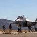 A military fifth-generation stealth fighter jet, identifiable as an F-35, is positioned on a tarmac or forward operating strip in a semi-arid, hilly environment. Ground crew personnel in combat gear are actively working around the aircraft, with at least one person handling what appears to be a fuel hose. The scene suggests a forward arming and refueling point (FARP) or expeditionary ground operations exercise.