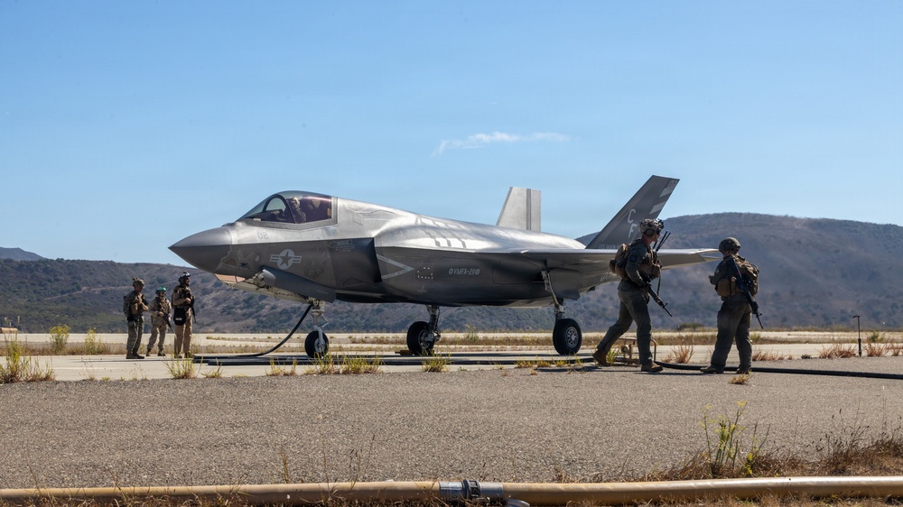 A U.S. military F-35 fighter jet is on a tarmac or forward operating strip, connected to a fuel hose, surrounded by several armed personnel in tactical gear. The aircraft bears U.S. Air Force/Marine Corps markings and appears to be undergoing a forward area refueling point (FARP) operation. The setting appears to be a semi-arid training or operational environment with hills visible in the background.