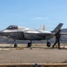A U.S. military F-35 fighter jet is on a tarmac or forward operating strip, connected to a fuel hose, surrounded by several armed personnel in tactical gear. The aircraft bears U.S. Air Force/Marine Corps markings and appears to be undergoing a forward area refueling point (FARP) operation. The setting appears to be a semi-arid training or operational environment with hills visible in the background.