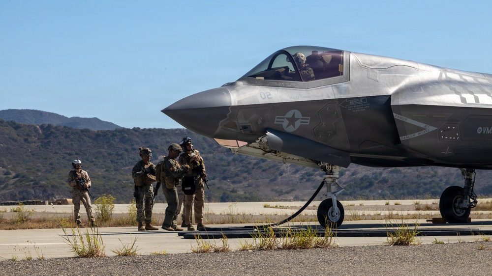 A U.S. Marine Corps F-35B Lightning II stealth fighter jet, marked with tail number '02' and a VMFA squadron designation, is parked on a tarmac in what appears to be a semi-arid training environment. Several military personnel in combat uniforms are gathered near the nose of the aircraft, while a pilot is visible in the cockpit. The setting suggests a forward operating base or expeditionary airfield exercise.
