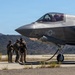A U.S. Marine Corps F-35B Lightning II stealth fighter jet, marked with tail number '02' and a VMFA squadron designation, is parked on a tarmac in what appears to be a semi-arid training environment. Several military personnel in combat uniforms are gathered near the nose of the aircraft, while a pilot is visible in the cockpit. The setting suggests a forward operating base or expeditionary airfield exercise.