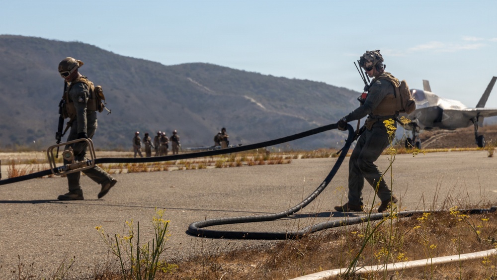Military personnel in full combat gear are conducting a forward arming and refueling point (FARP) operation on a tarmac in a dry, hilly terrain. Two foreground personnel are handling large fuel hoses, while additional troops are visible in the background near what appears to be an F-35 fighter jet. The setting suggests a field exercise or expeditionary operation at a temporary or austere airstrip.