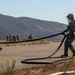Military personnel in full combat gear are conducting a forward arming and refueling point (FARP) operation on a tarmac in a dry, hilly terrain. Two foreground personnel are handling large fuel hoses, while additional troops are visible in the background near what appears to be an F-35 fighter jet. The setting suggests a field exercise or expeditionary operation at a temporary or austere airstrip.