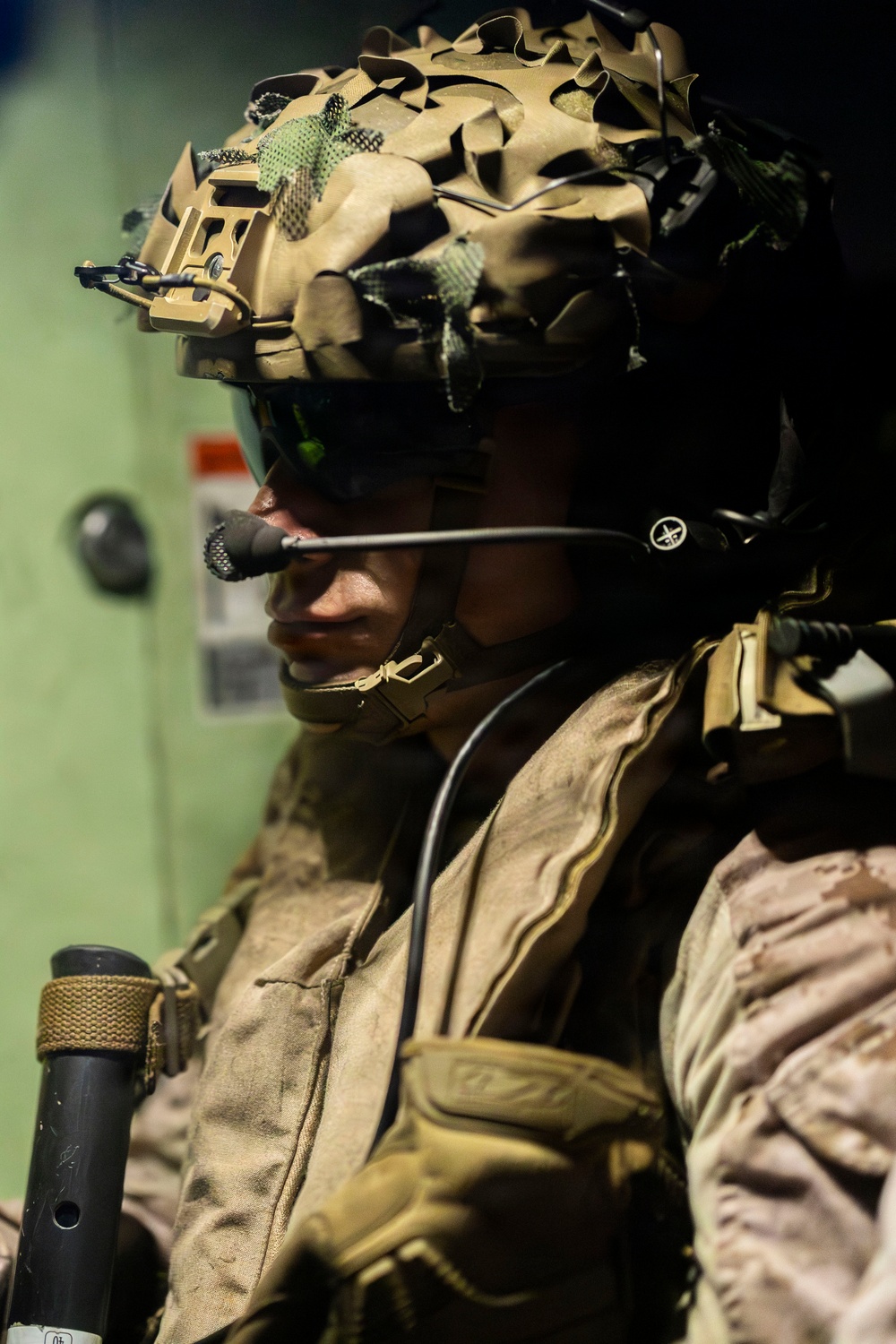 A close-up portrait of a military service member in full tactical gear, photographed in profile/three-quarter view. The individual is equipped with advanced communications equipment and a camouflage-covered combat helmet. The image appears to be taken inside a military vehicle or vessel based on the green metal background.