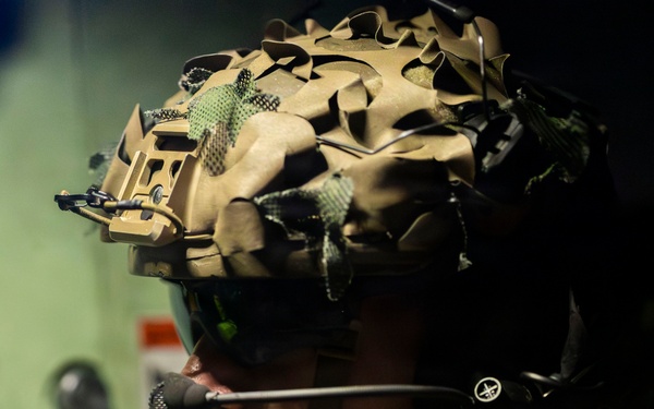 A close-up portrait of a military service member in full tactical gear, photographed in profile/three-quarter view. The individual is equipped with advanced communications equipment and a camouflage-covered combat helmet. The image appears to be taken inside a military vehicle or vessel based on the green metal background.