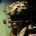 A close-up portrait of a military service member in full tactical gear, photographed in profile/three-quarter view. The individual is equipped with advanced communications equipment and a camouflage-covered combat helmet. The image appears to be taken inside a military vehicle or vessel based on the green metal background.