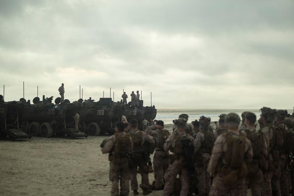 A large group of military personnel in camouflage uniforms are assembled on a sandy beach alongside multiple armored amphibious vehicles. The scene appears to depict a military amphibious landing exercise or operation, with soldiers gathered near the waterline. The overcast sky and muted lighting give the image a somber, operational atmosphere.