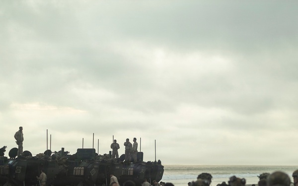 A large group of military personnel in camouflage uniforms are assembled on a sandy beach alongside multiple armored amphibious vehicles. The scene appears to depict a military amphibious landing exercise or operation, with soldiers gathered near the waterline. The overcast sky and muted lighting give the image a somber, operational atmosphere.