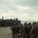 A large group of military personnel in camouflage uniforms are assembled on a sandy beach alongside multiple armored amphibious vehicles. The scene appears to depict a military amphibious landing exercise or operation, with soldiers gathered near the waterline. The overcast sky and muted lighting give the image a somber, operational atmosphere.