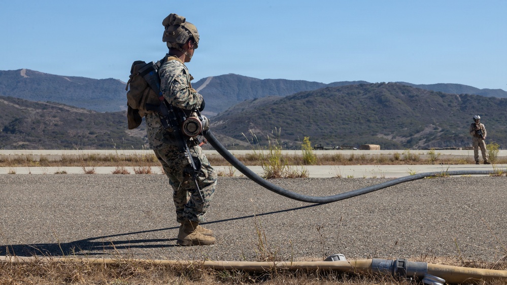 A military service member in camouflage uniform and combat helmet stands on a paved surface holding a large black fuel or fluid transfer hose. A second uniformed personnel is visible in the background. The setting appears to be a military airfield or training area with arid terrain and mountains in the background.