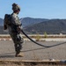 A military service member in camouflage uniform and combat helmet stands on a paved surface holding a large black fuel or fluid transfer hose. A second uniformed personnel is visible in the background. The setting appears to be a military airfield or training area with arid terrain and mountains in the background.