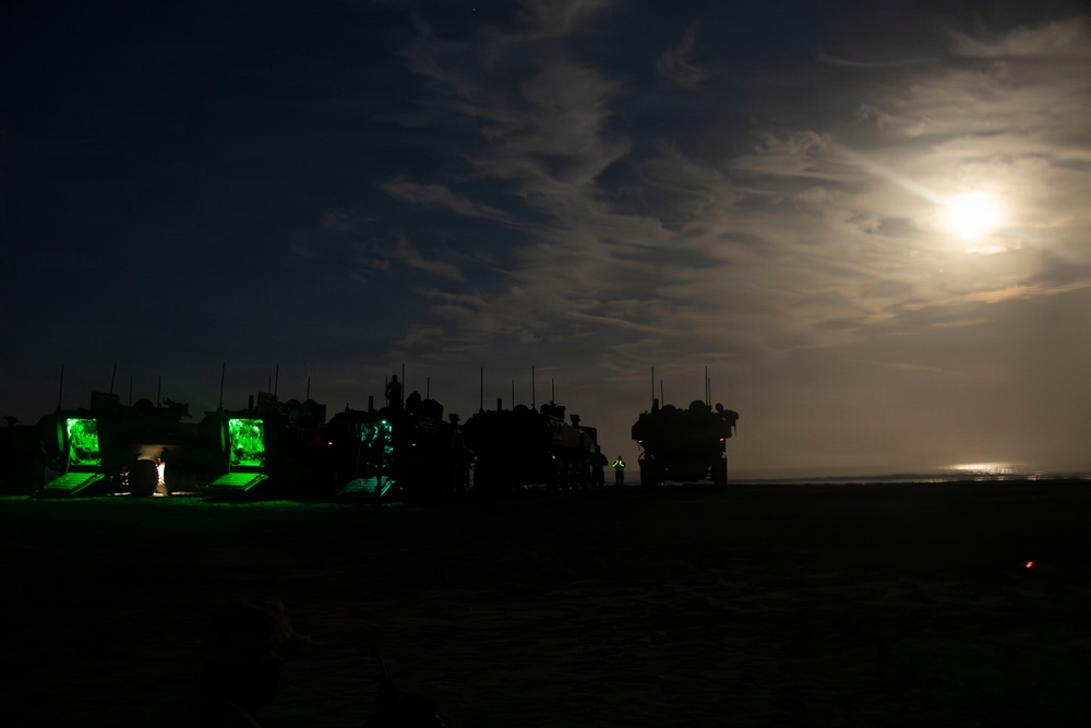 A nighttime military scene showing a convoy or staging area of military vehicles silhouetted against a moonlit sky. Several vehicles have open compartments illuminated with bright green interior lighting, suggesting active operations or equipment checks. A full or near-full moon partially obscured by clouds provides ambient illumination over what appears to be a flat, arid terrain.