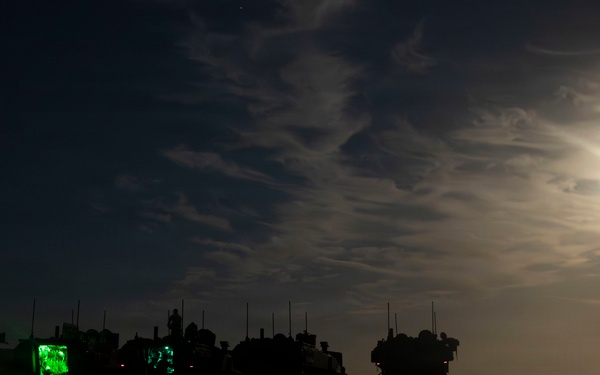 A nighttime military scene showing a convoy or staging area of military vehicles silhouetted against a moonlit sky. Several vehicles have open compartments illuminated with bright green interior lighting, suggesting active operations or equipment checks. A full or near-full moon partially obscured by clouds provides ambient illumination over what appears to be a flat, arid terrain.