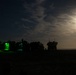 A nighttime military scene showing a convoy or staging area of military vehicles silhouetted against a moonlit sky. Several vehicles have open compartments illuminated with bright green interior lighting, suggesting active operations or equipment checks. A full or near-full moon partially obscured by clouds provides ambient illumination over what appears to be a flat, arid terrain.