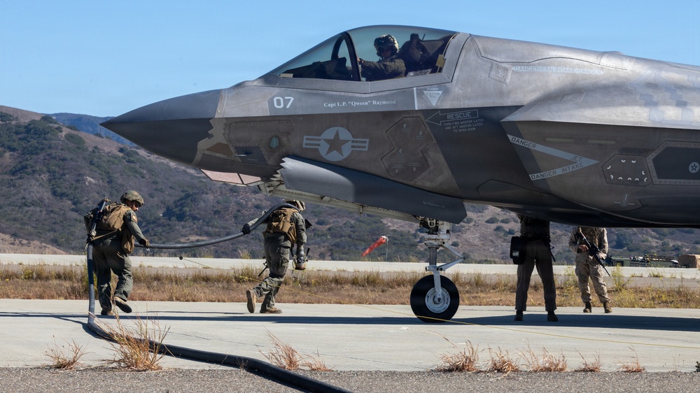 A U.S. military F-35 stealth fighter jet (tail number 07, assigned to 'Capt L.P. Qween Raymann') is on a tarmac with ground crew personnel conducting what appears to be a forward arming and refueling point (FARP) operation. Multiple personnel in tactical gear are working around the aircraft, with one handling a fuel hose. The setting appears to be a military airfield with hills and dry vegetation visible in the background.