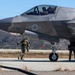 A U.S. military F-35 stealth fighter jet (tail number 07, assigned to 'Capt L.P. Qween Raymann') is on a tarmac with ground crew personnel conducting what appears to be a forward arming and refueling point (FARP) operation. Multiple personnel in tactical gear are working around the aircraft, with one handling a fuel hose. The setting appears to be a military airfield with hills and dry vegetation visible in the background.