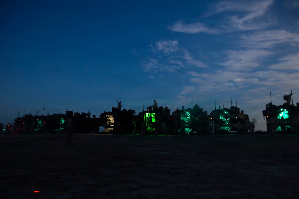 A nighttime military scene on what appears to be a beach or sandy terrain, showing a line of military vehicles illuminated by green interior lighting. Multiple silhouetted personnel are visible moving around the vehicles. The scene suggests a military amphibious or beach landing operation conducted under low-light conditions.