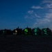 A nighttime military scene on what appears to be a beach or sandy terrain, showing a line of military vehicles illuminated by green interior lighting. Multiple silhouetted personnel are visible moving around the vehicles. The scene suggests a military amphibious or beach landing operation conducted under low-light conditions.
